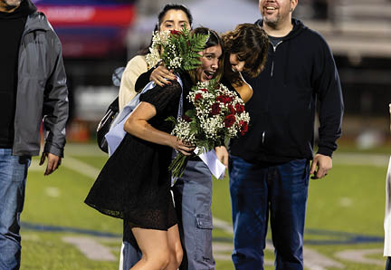 A person in a black dress holding a bouquet of flowers stands on a sports field next to two people wearing casual clothing. Description generated by AI