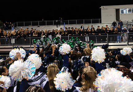 A large group of people in sports uniforms and cheerleading outfits are gathered on a field at night with some holding pom-poms and helmets raised, while a crowd watches from bleachers in the background. Description generated by AI