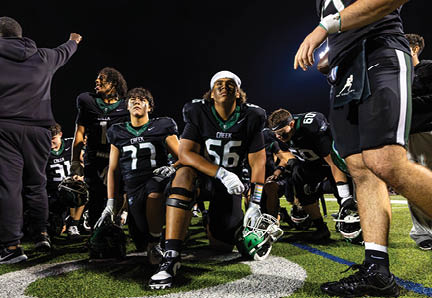 A group of football players in black uniforms are gathered on a field at night, some kneeling and others standing, with one player holding a helmet. Description generated by AI
