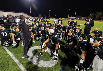 A group of football players in black uniforms are gathered on a field at night listening to a coach who is standing and speaking to them. Description generated by AI