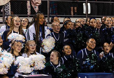 A group of people in cheerleading uniforms are smiling and holding pom-poms while standing closely together in a stadium setting. Description generated by AI