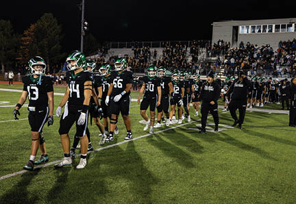 A group of people wearing black sports uniforms and helmets walk together on a football field at night with spectators in the background. Description generated by AI