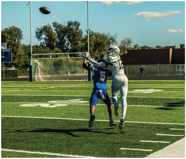 Two people in football uniforms are jumping to catch a football on a sunny field with a goalpost and scoreboard in the background. Description generated by AI