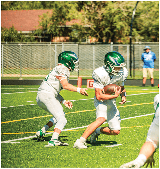 Two people in green helmets and white football uniforms are playing on a football field with one holding the ball and the other reaching out to receive or block. Description generated by AI