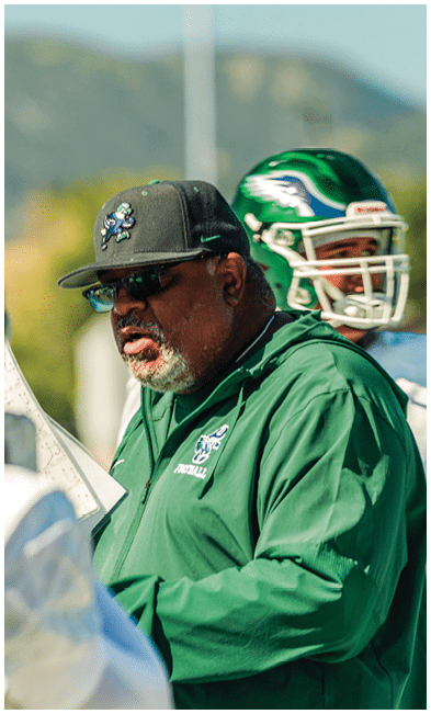 A person wearing a green football helmet and white jersey with a small American flag on the back is facing another person holding a play sheet outdoors with a blurred natural background. Description generated by AI