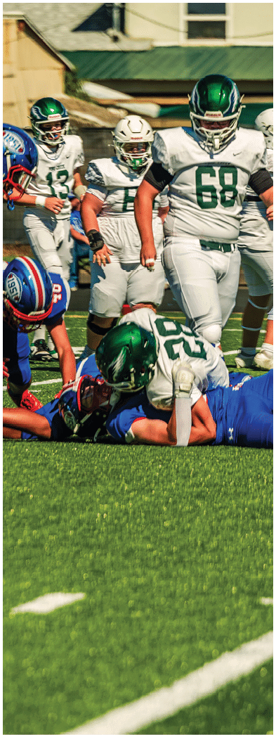 Two people in blue sports uniforms are engaged in a physical struggle on a green artificial turf field near a fence and buildings. Description generated by AI