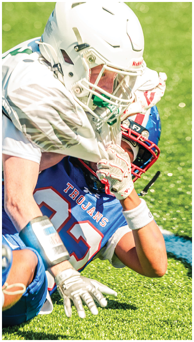 Two people wearing sports uniforms are engaged in a tackle on a grassy field during a game. Description generated by AI