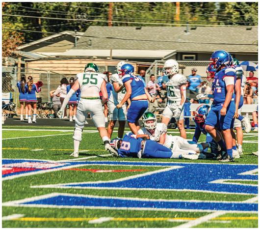 Players in blue and white football uniforms are engaged in a play on a green field with cheerleaders and spectators in the background. Description generated by AI