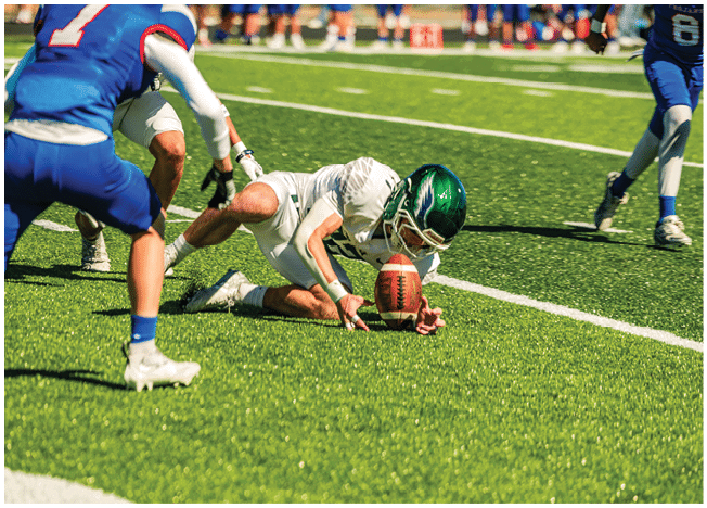 A person in a white football uniform and green helmet is on the ground reaching for a football while two people in blue uniforms approach on a sunny football field. Description generated by AI