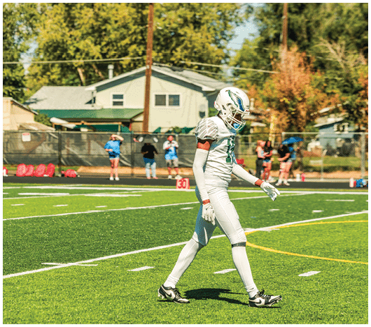 A person in a white football uniform and helmet is walking on a green football field with a few people standing in the background near a fence and houses. Description generated by AI