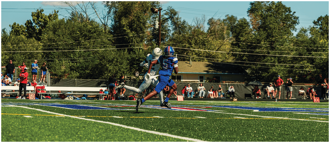 Two people in football uniforms are jumping and reaching for a football in an outdoor field with trees and spectators in the background. Description generated by AI