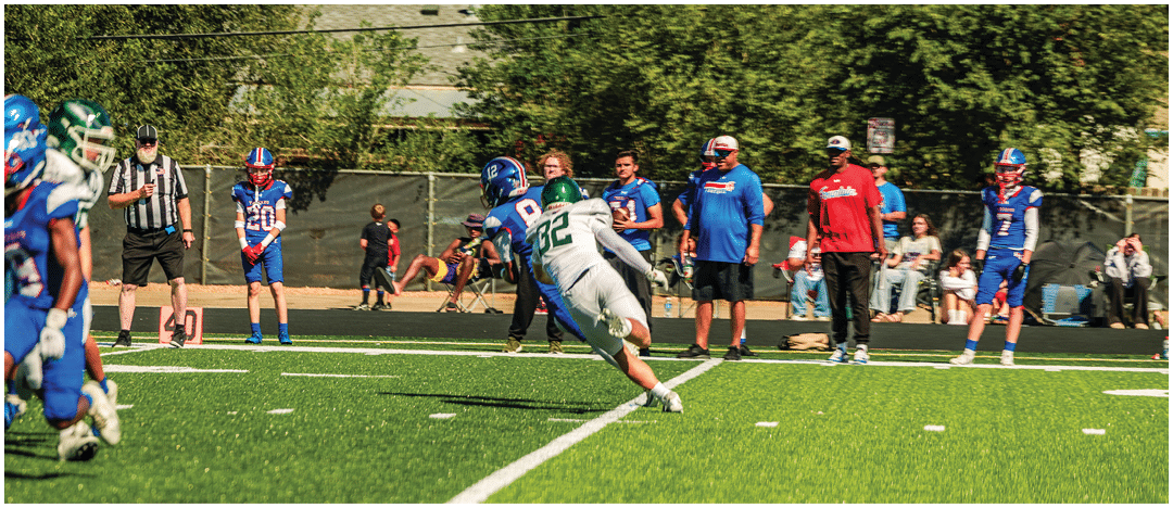 A person in a white football uniform is diving to catch a football while several people in blue uniforms and casual clothing watch from the sidelines on a sunny day. Description generated by AI