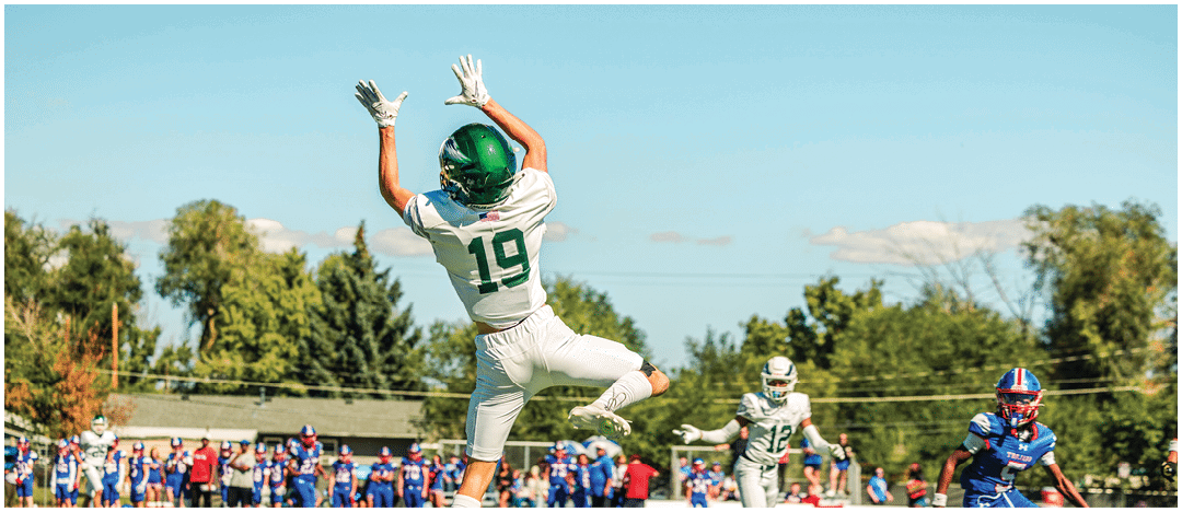 A person wearing a green helmet and white sports uniform with the number 19 is jumping to catch a football during a game on a sunny day with other players and spectators in the background. Description generated by AI