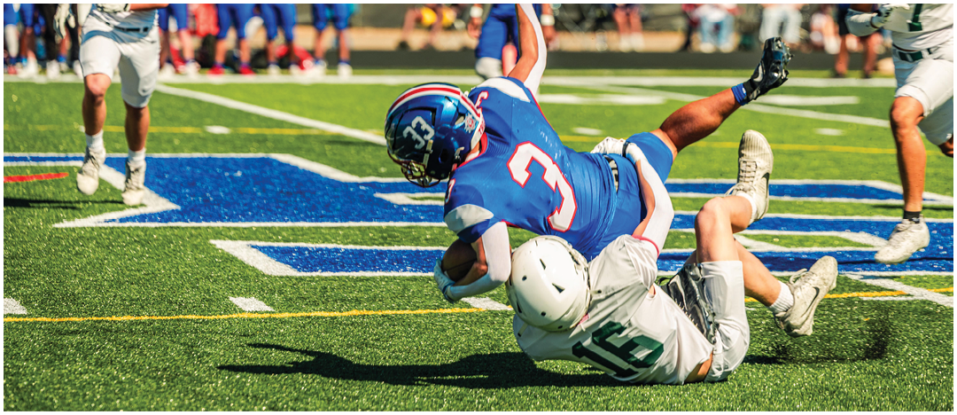 A person in blue football gear is falling backward on a field while two people in white and green uniforms approach from either side. Description generated by AI