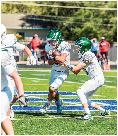 People in white football uniforms are playing on a green field with one person holding the ball and running while others follow. Description generated by AI