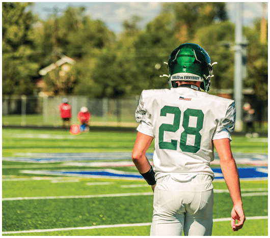 A person wearing a green helmet and white football uniform with the number 28 stands on a football field facing away from the camera. Description generated by AI