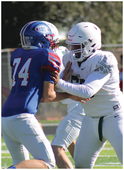 Two people in white football uniforms and helmets are engaged in a play on a football field with a person in casual clothing walking in the background. Description generated by AI