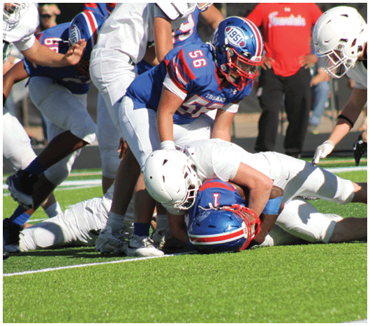 Two people in football uniforms are on the ground with one tackling the other while other players stand nearby on a green field. Description generated by AI
