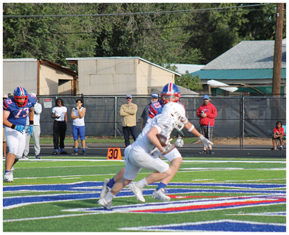 Two people in football uniforms are running on a field with a football while several people watch from the sidelines. Description generated by AI