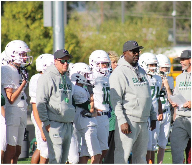 A group of people wearing football helmets and gray hoodies stand together on a sports field with trees in the background. Description generated by AI