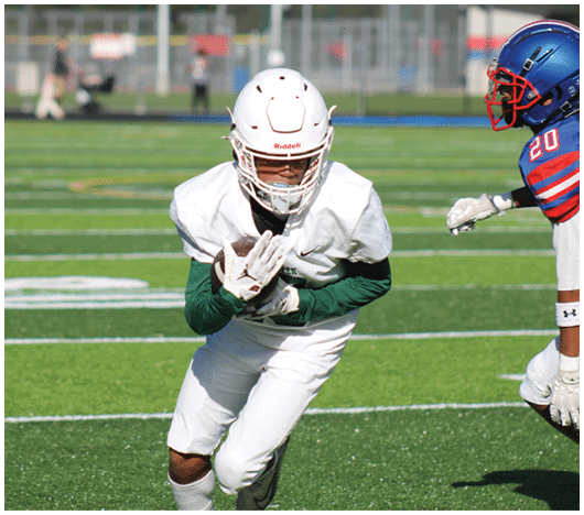 A person in white football gear is running on a green field while another person in blue and red football gear approaches from the side. Description generated by AI