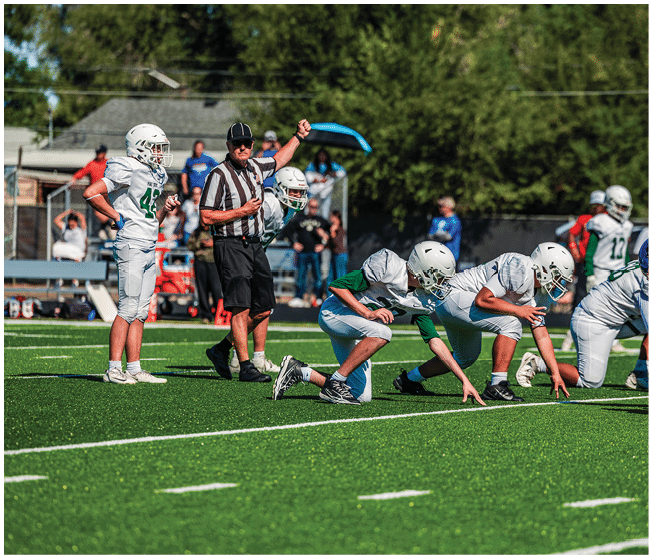Two people wearing football uniforms are on a field with one person in a green jersey running towards the other person in a white jersey against a backdrop of mountains and a partly cloudy sky. Description generated by AI