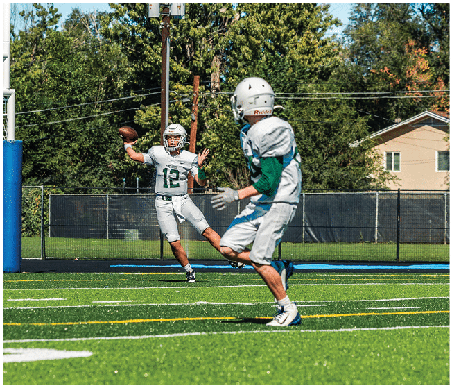 Two people wearing football uniforms are on a field with one person in a green jersey running towards the other person in a white jersey against a backdrop of mountains and a partly cloudy sky. Description generated by AI