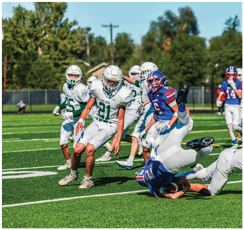 Two people wearing football uniforms are on a field with one person in a green jersey running towards the other person in a white jersey against a backdrop of mountains and a partly cloudy sky. Description generated by AI