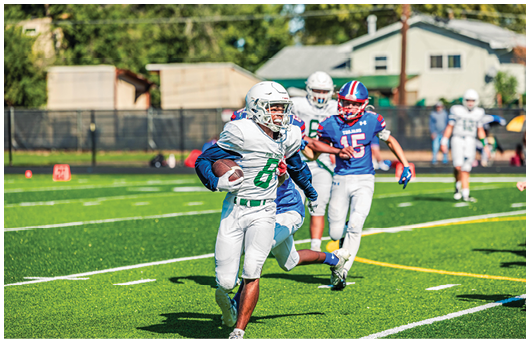 Two people wearing football uniforms are on a field with one person in a green jersey running towards the other person in a white jersey against a backdrop of mountains and a partly cloudy sky. Description generated by AI