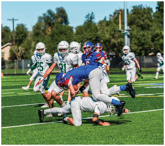 Five people in football uniforms stand on a field facing three other people in green and white football uniforms with helmets under a blue sky. Description generated by AI