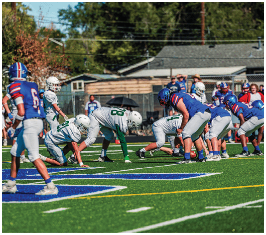 Five people in football uniforms stand on a field facing three other people in green and white football uniforms with helmets under a blue sky. Description generated by AI