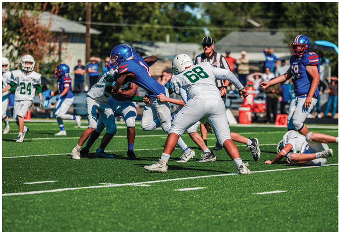 Five people in football uniforms stand on a field facing three other people in green and white football uniforms with helmets under a blue sky. Description generated by AI