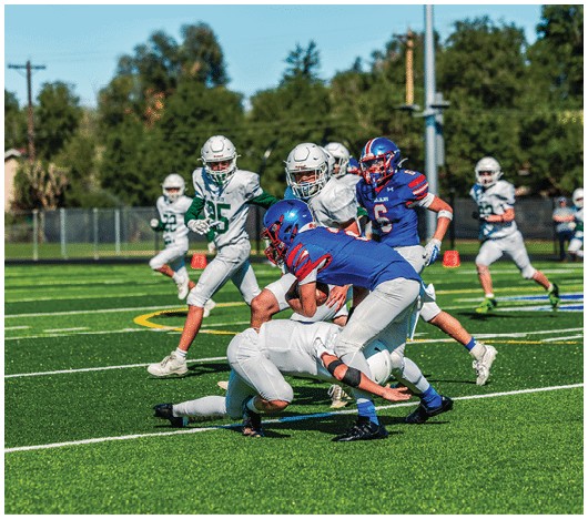 Five people in football uniforms stand on a field facing three other people in green and white football uniforms with helmets under a blue sky. Description generated by AI