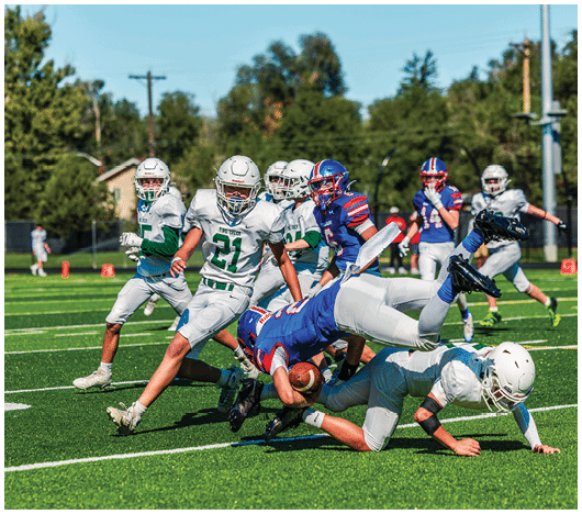 Five people in football uniforms stand on a field facing three other people in green and white football uniforms with helmets under a blue sky. Description generated by AI