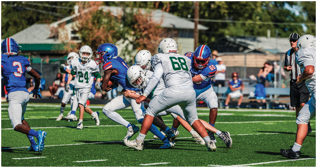 Five people in football uniforms stand on a field facing three other people in green and white football uniforms with helmets under a blue sky. Description generated by AI