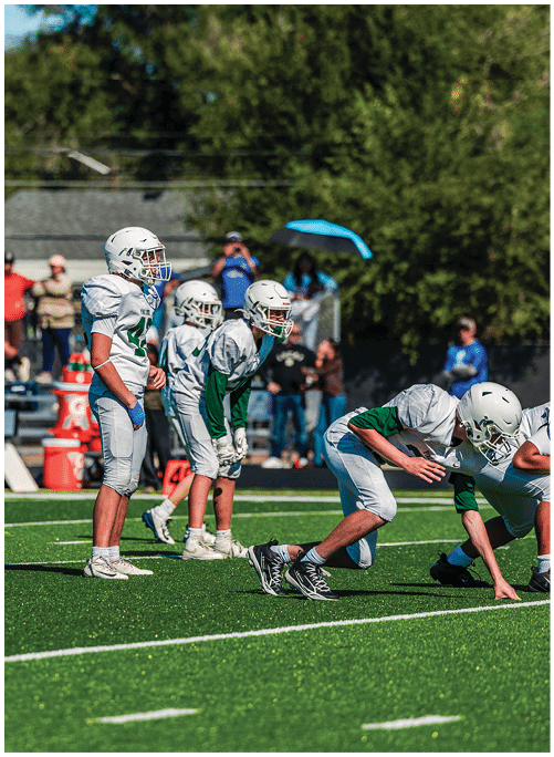 Five people in football uniforms stand on a field facing three other people in green and white football uniforms with helmets under a blue sky. Description generated by AI
