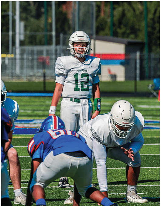 Five people in football uniforms stand on a field facing three other people in green and white football uniforms with helmets under a blue sky. Description generated by AI