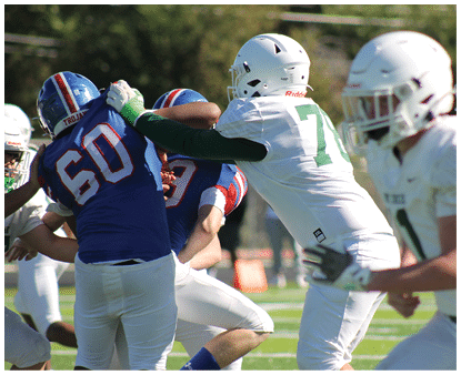 People wearing football helmets and uniforms are engaged in a play on a grassy field during a game. Description generated by AI