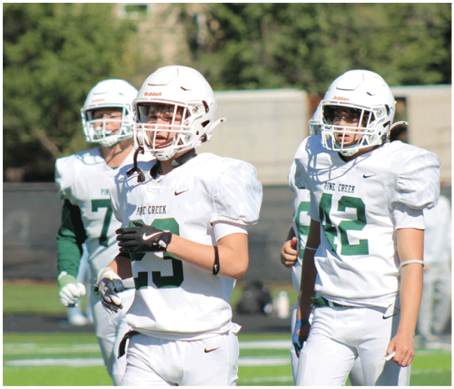 Three people wearing white football uniforms and helmets are standing on a green field with trees and a fence in the background. Description generated by AI
