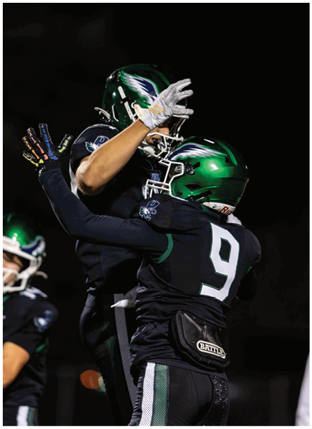Two people wearing green helmets and black football uniforms celebrate by embracing on a field at night. Description generated by AI