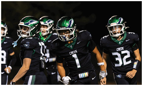 People wearing black football uniforms with green helmets are on a field, appearing to be in motion during a game or practice. Description generated by AI