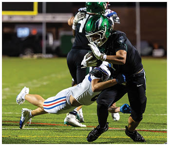 A person in a black uniform and green helmet is running with a football while another person in a white uniform and blue helmet is diving to tackle them on a lit football field at night. Description generated by AI