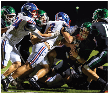 A group of football players in white and black uniforms are engaged in a tackle during a nighttime game on a grassy field. Description generated by AI