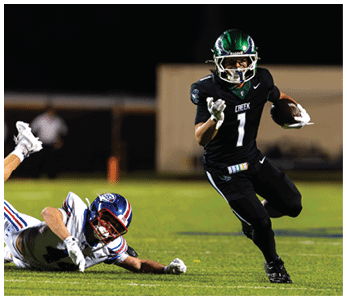 A person in a black football uniform with the number 7 is running with the ball while another person in a white football uniform is falling on the grass field behind them. Description generated by AI