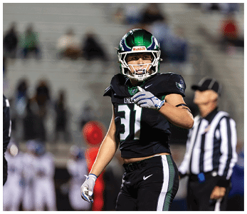 A person wearing a black football uniform with the number 31 and a green helmet is standing on a field with blurred people and a referee in the background. Description generated by AI