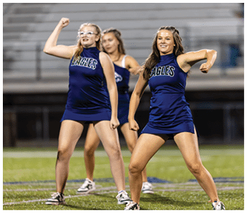 Three adults in matching blue cheerleading uniforms with the word Eagles perform a synchronized routine on a sports field with empty bleachers in the background. Description generated by AI
