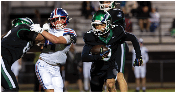 A person in a black football uniform is running with the ball while being pursued by a person in a white football uniform and another person in a black football uniform on a field. Description generated by AI