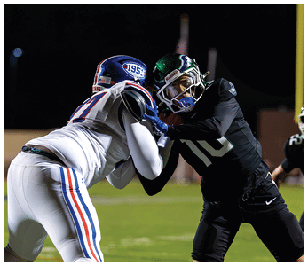 Two people in football uniforms are engaged in a physical block on a field during a nighttime game. Description generated by AI