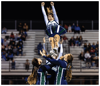 A person in a cheerleading uniform with “EAGLES" on the top is being lifted into the air by several hands during a nighttime event. Description generated by AI