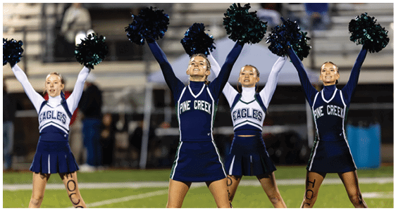 Four cheerleaders in blue and white uniforms with pom-poms raised are performing on a football field at night. Description generated by AI